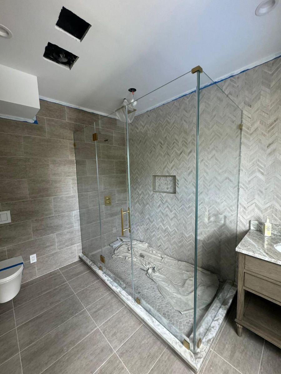 A modern bathroom featuring a glass walk-in shower with light gray herringbone tile, neutral flooring, and a vanity.
