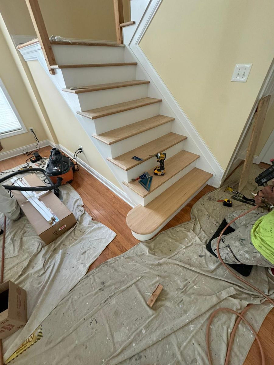 A worker renovates wooden stairs in a home, with tools, building supplies, and drop cloths covering the floor.