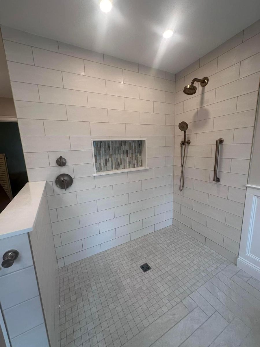 A modern walk-in shower with white brick-style wall tiles, a recessed shelf, bronze fixtures, and light stone flooring.
