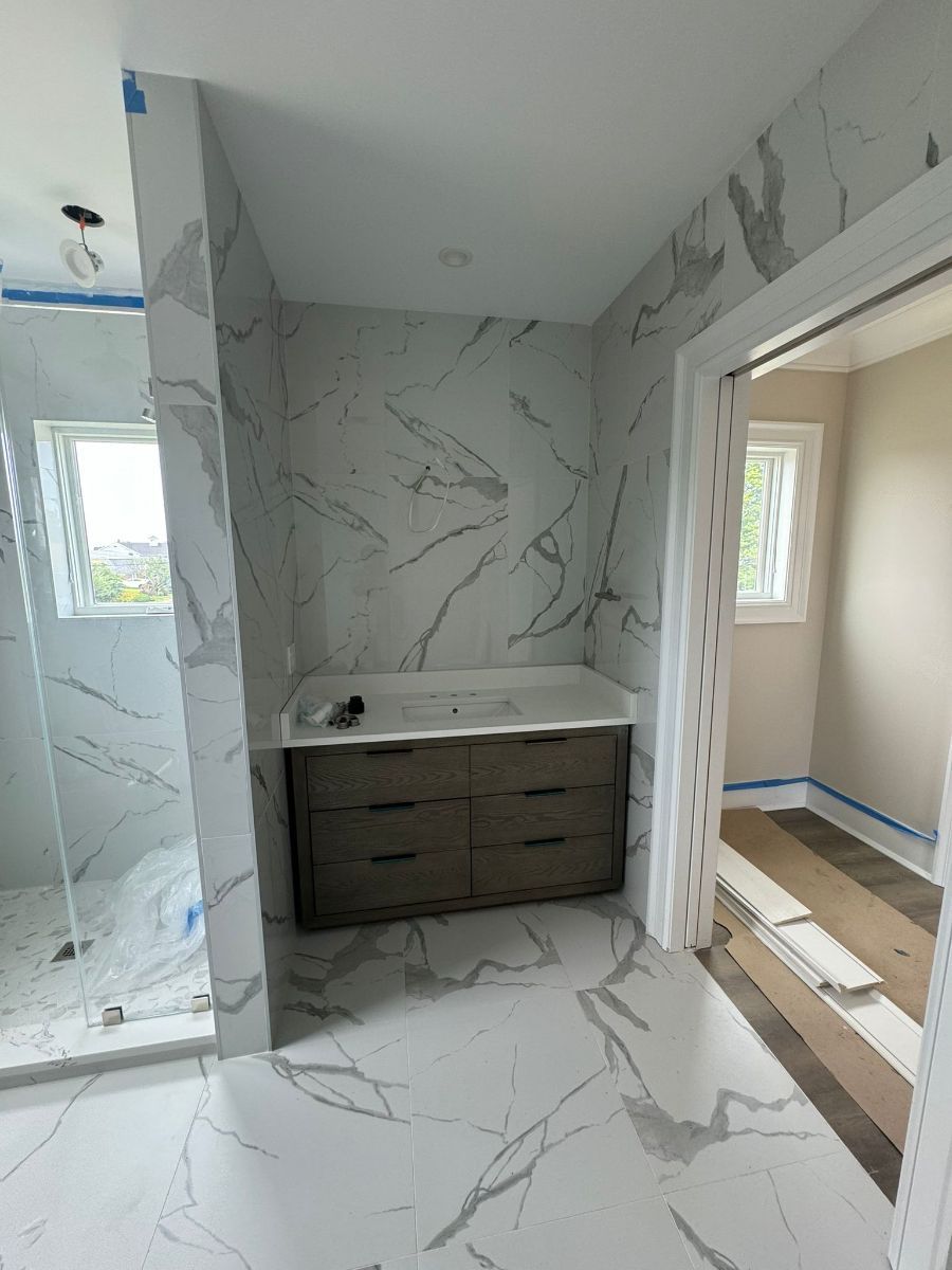 A bathroom vanity with dark wood drawers against a white marble-tiled wall, flanked by a glass-walled shower and doorway.