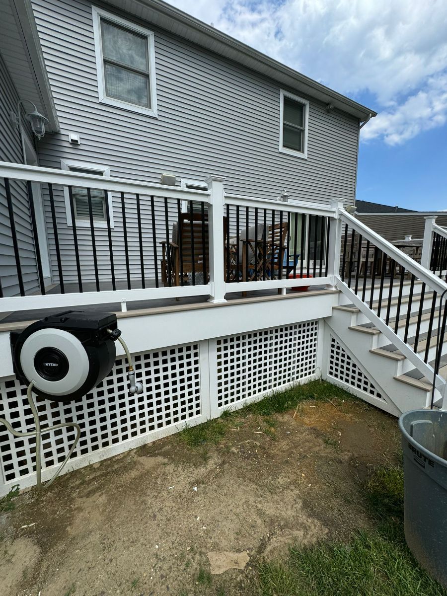 A grey-sided house features a white deck with black balusters, lattice skirting, stairs, and a hose reel attached below.