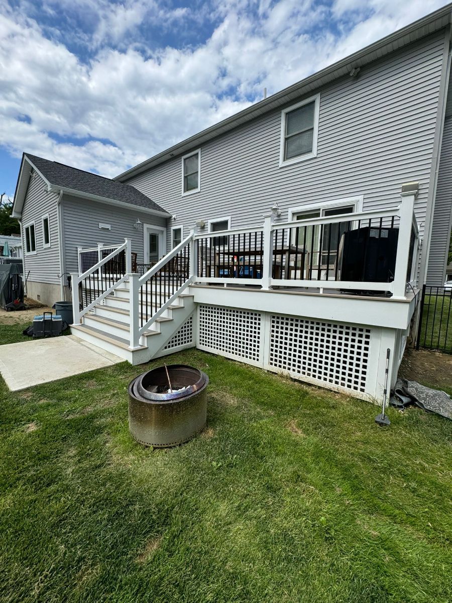 A multi-level gray house with a white deck, stairs, lattice skirting, and a concrete fire pit in the grassy backyard.