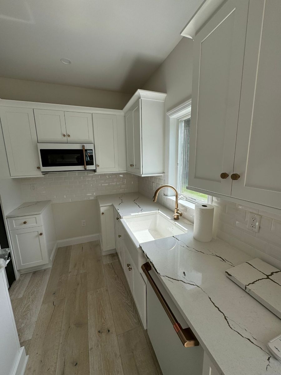 A bright, modern kitchen with white cabinets, light wood floors, and a white farmhouse sink with a gold faucet.