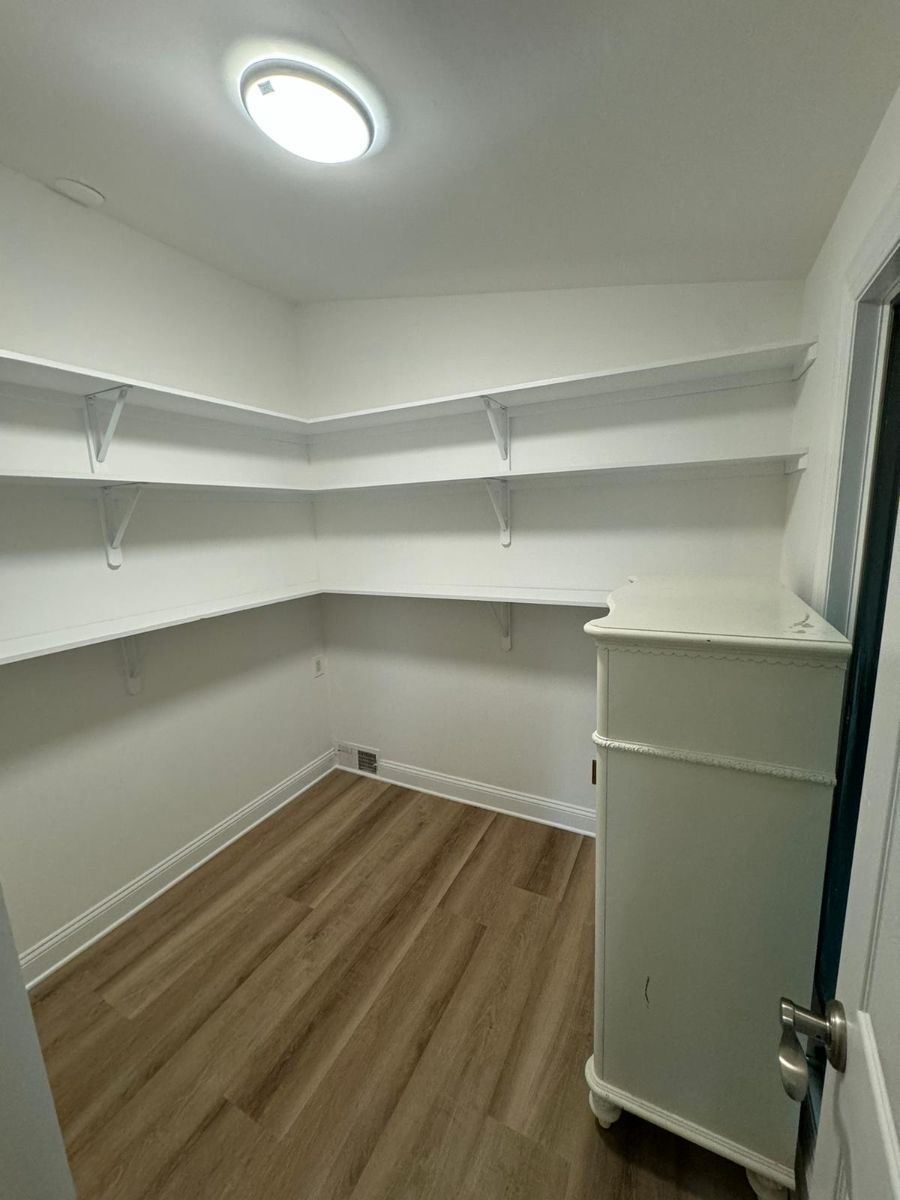 An empty pantry featuring white L-shaped shelving, white walls, light wood-look flooring, and a ceiling light.