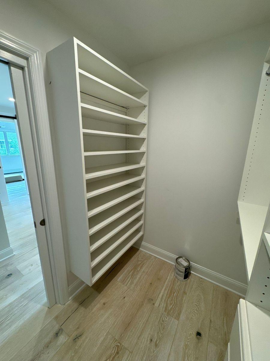 A white custom shoe rack with multiple horizontal shelves mounted on a wall in a light-colored walk-in closet.