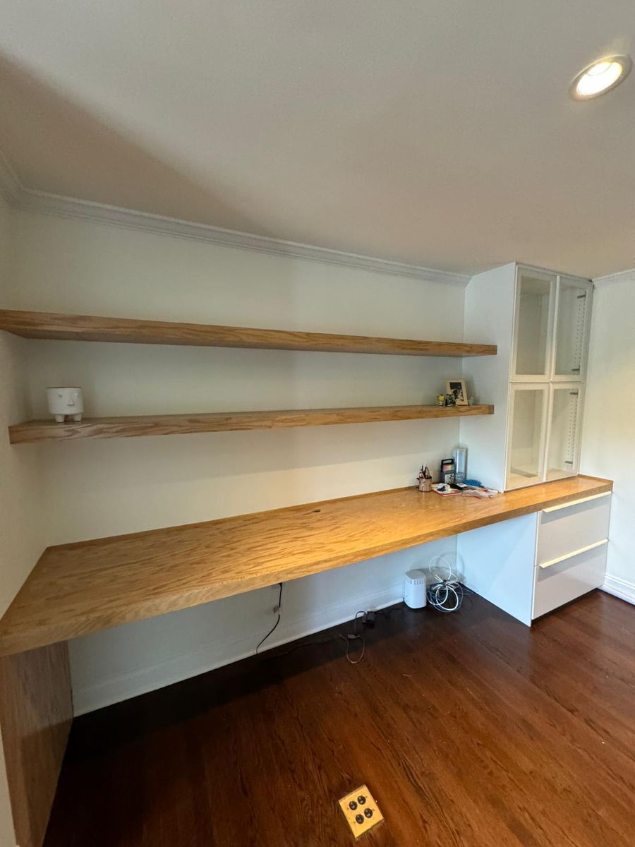 A room with light wood floating shelves above a long wooden desk, connected to a white cabinet against a white wall.