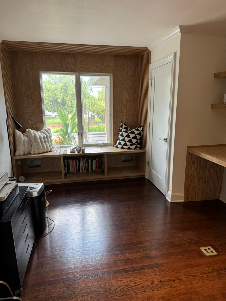 A window nook with wooden shelving, a white cushion, throw pillows, and a built-in desk in a room with dark wood floors.