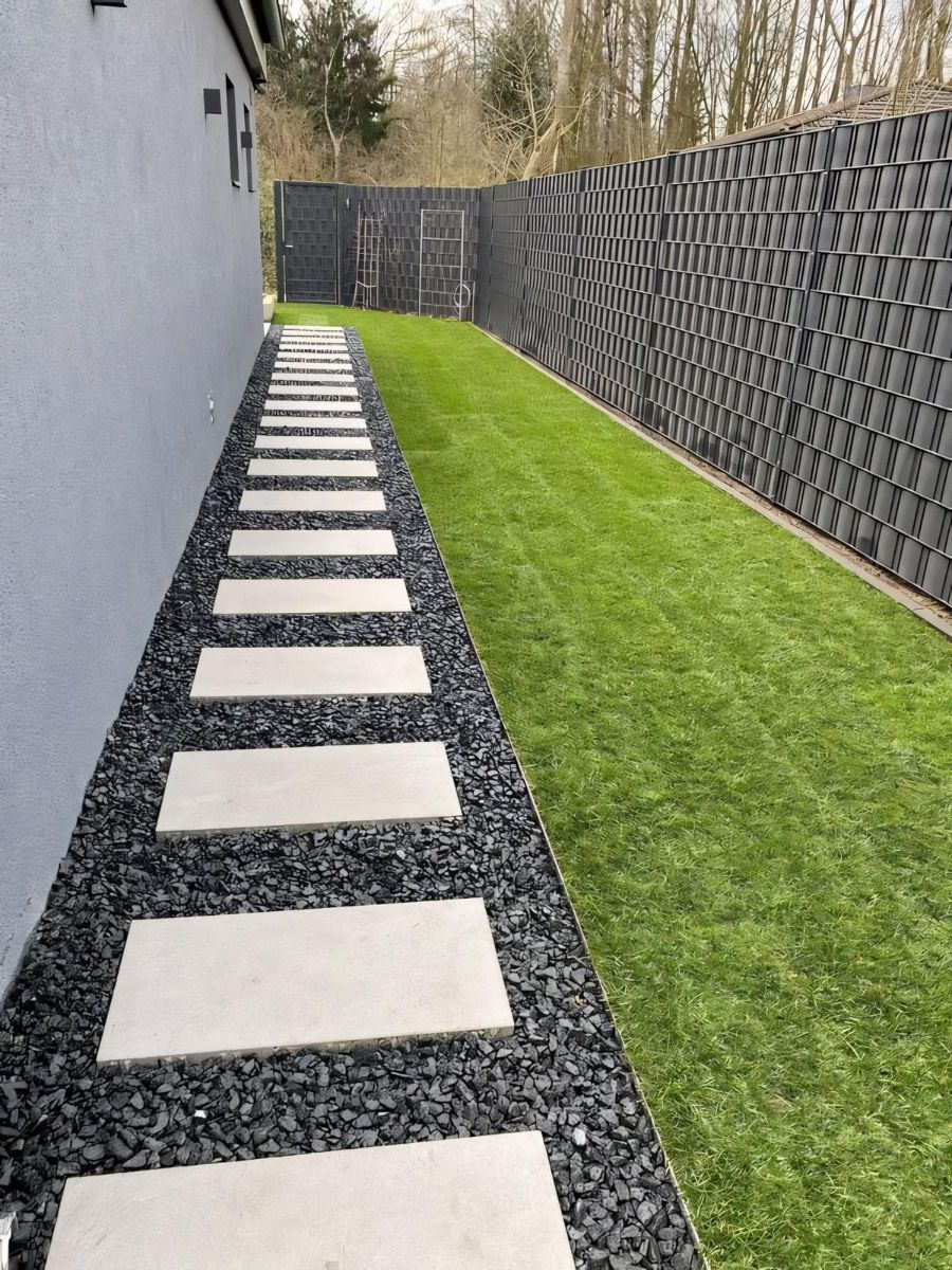 A side yard path with white rectangular pavers set in dark grey gravel next to a green lawn and a dark modern fence.
