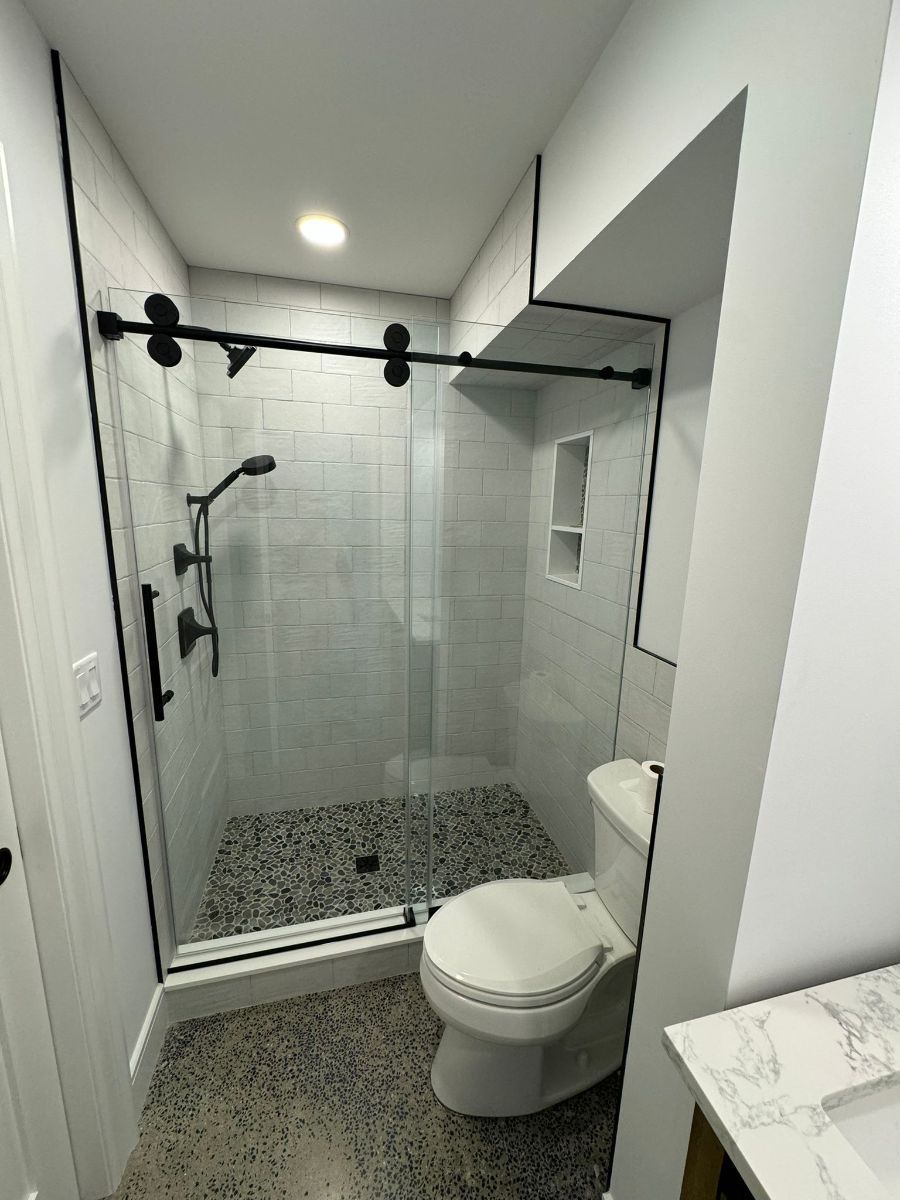 A modern bathroom with a glass-enclosed shower featuring a black track, pebble flooring, and a white toilet nearby.