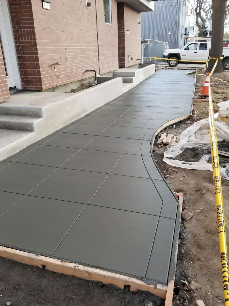 A freshly poured concrete sidewalk with square-grid scoring runs along the front of a brick building.