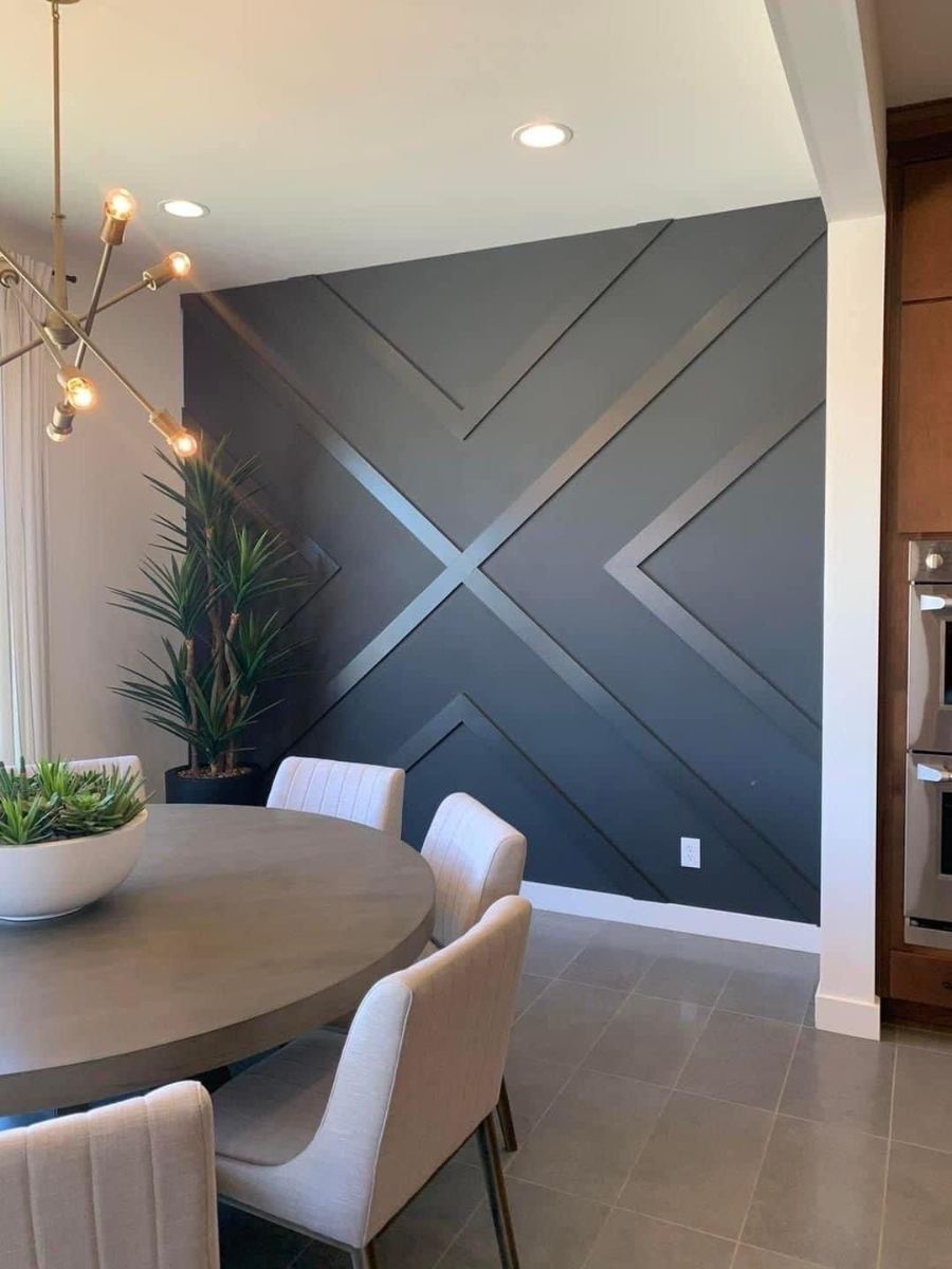 A dining area with a round gray table, light chairs, a potted plant, and a dark accent wall with geometric wood molding.