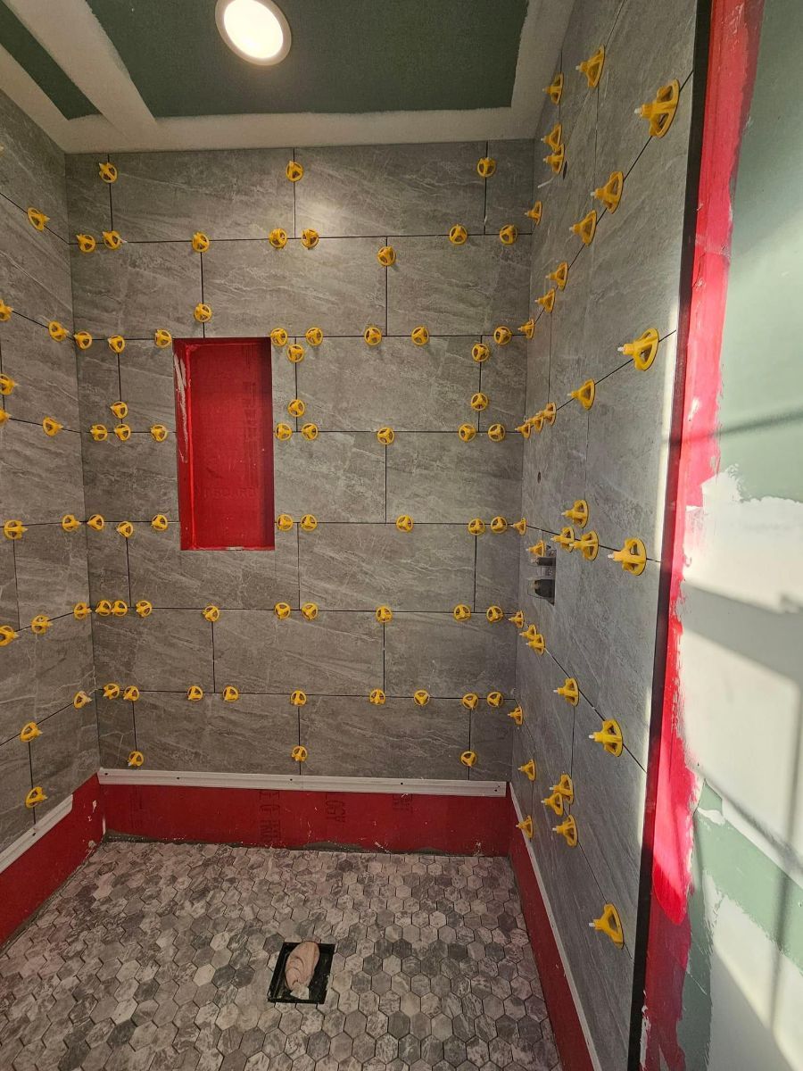 A shower stall under construction with gray stone wall tiles, yellow leveling clips, a red recessed niche, and stone floor.