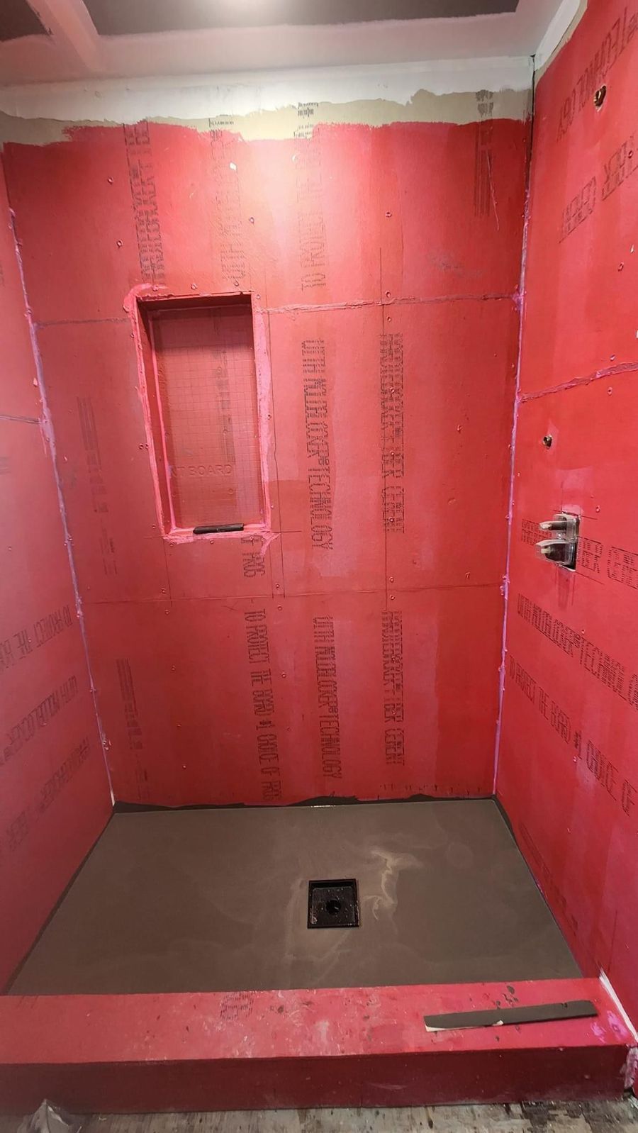 Shower alcove with red waterproof wall boards, a recessed niche, a mudded floor with a central drain, and exposed plumbing.