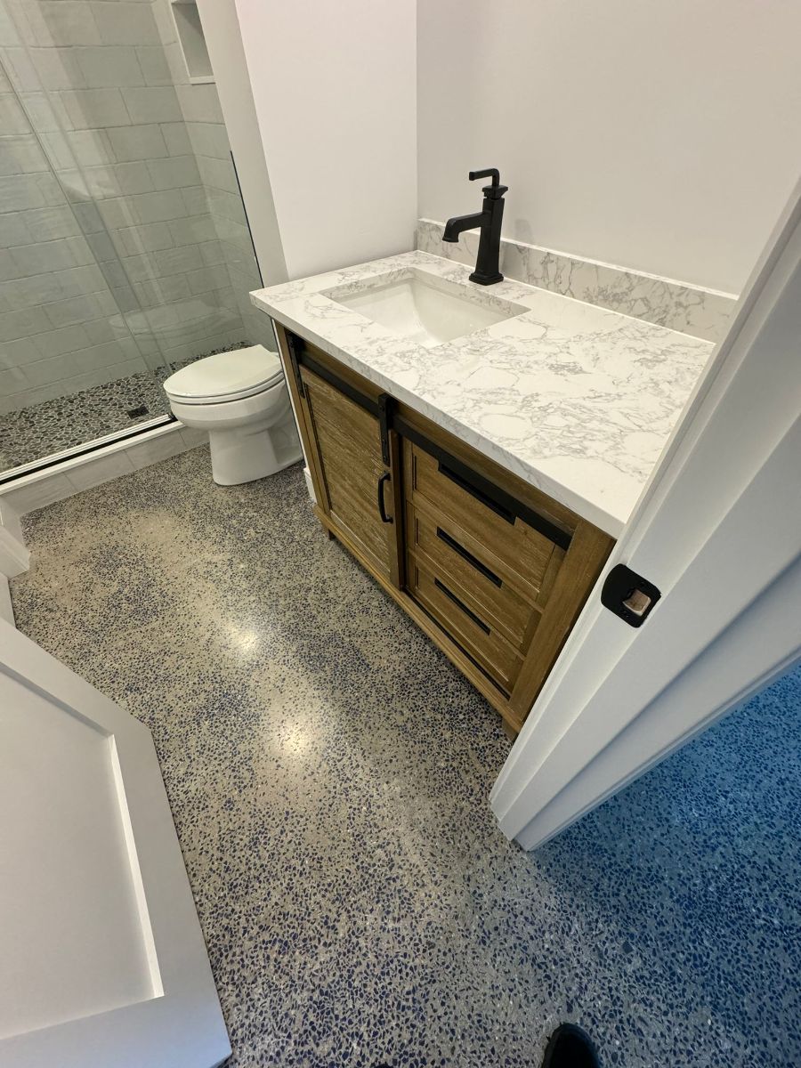 Modern bathroom with a wooden vanity, marble countertop, black faucet, tiled shower, and speckled flooring.