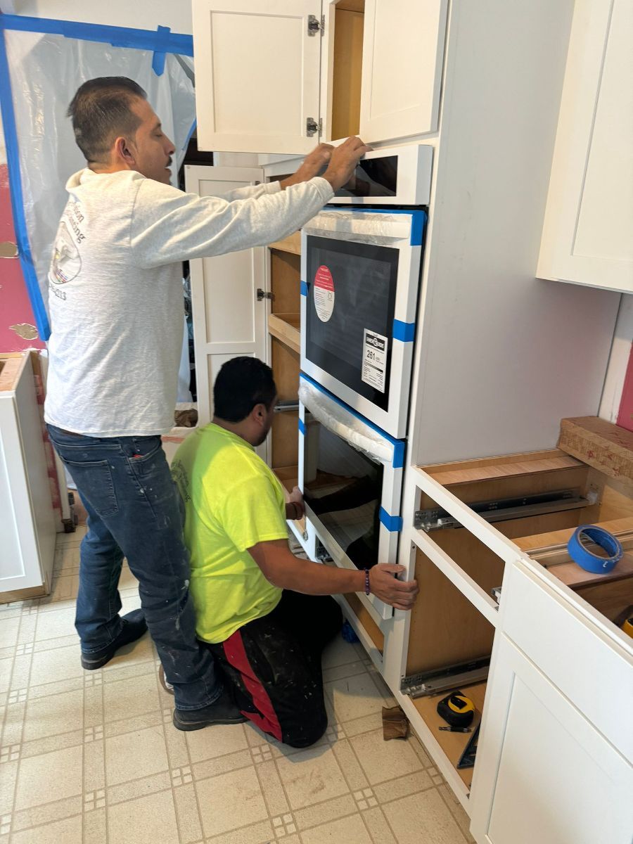 Two workers in a kitchen install a built-in double oven into a cabinet frame.