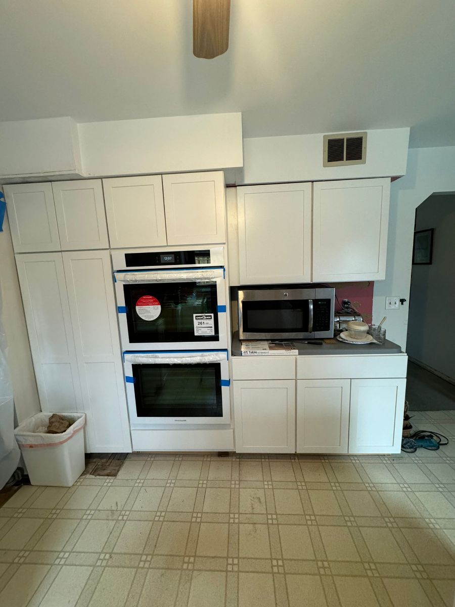 A kitchen wall featuring white cabinets, a double oven, a built-in microwave, and a small trash bin on a tiled floor.