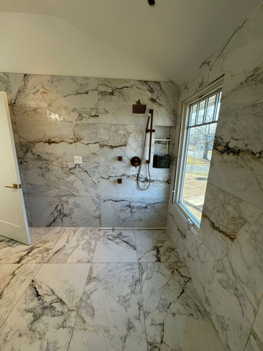 A walk-in shower with floor-to-ceiling white marble-patterned tiles, a handheld showerhead, and a rectangular window.