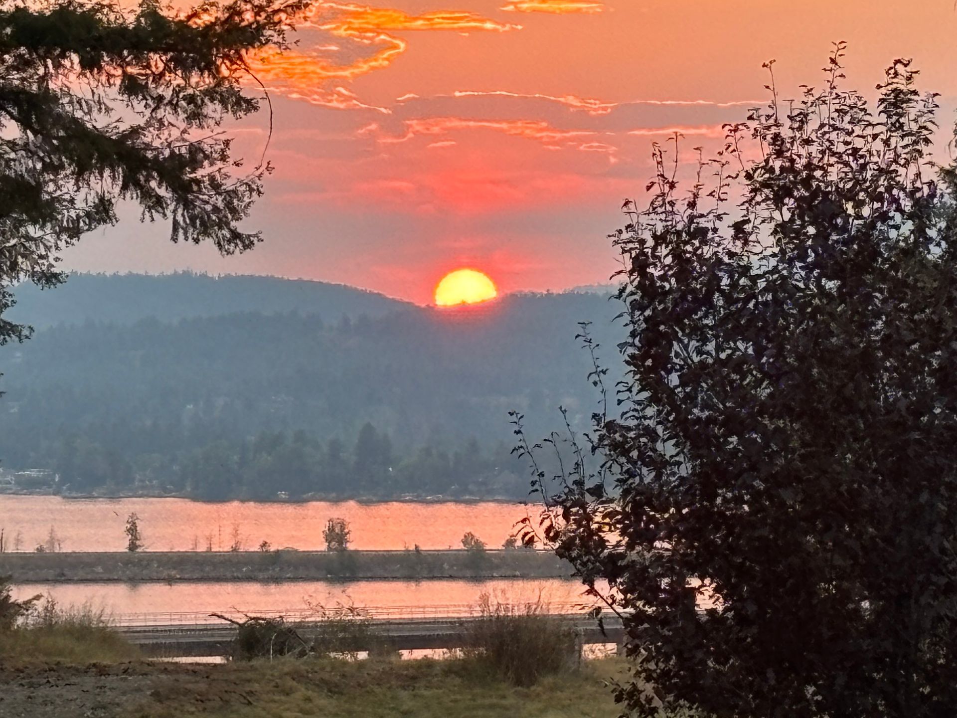 Sunset over a lake and hills, with orange sun, and silhouettes of trees and land.