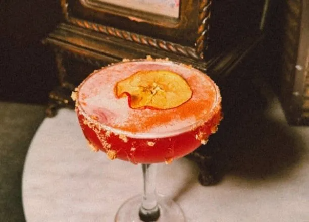 Red cocktail with apple slice garnish and sugared rim on a table.