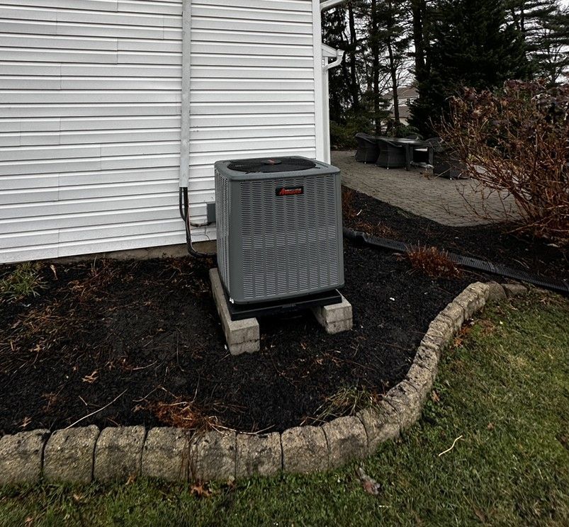 Outdoor air conditioning unit beside a white house, bordered by a stone-edged mulch bed.