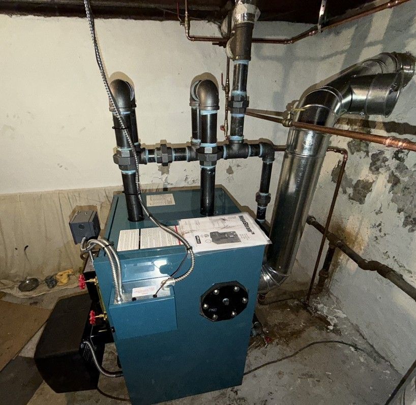 Boiler in a basement with exposed pipes, ducts, and a blue heating unit against a concrete wall.