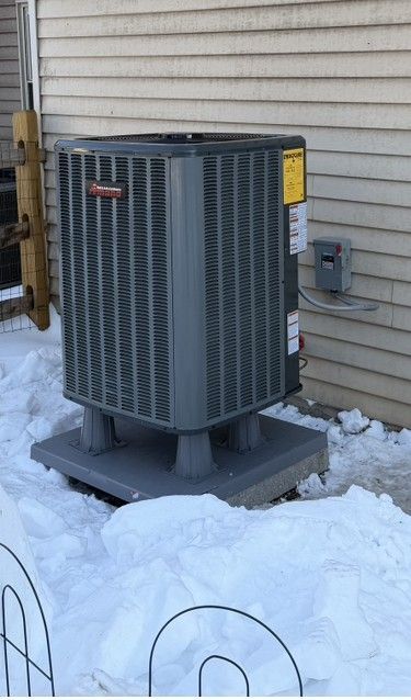 Outdoor air conditioner unit on a snowy ground beside a house wall