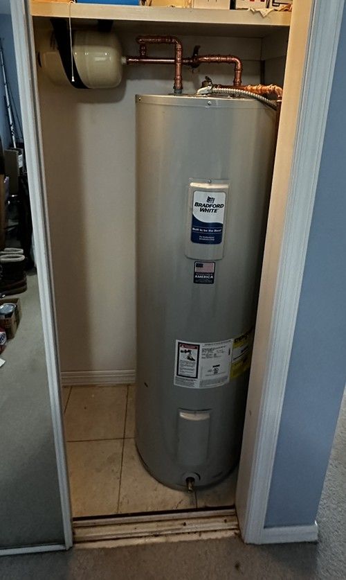 Water heater in a closet, with exposed pipes and a small utility tank above it.