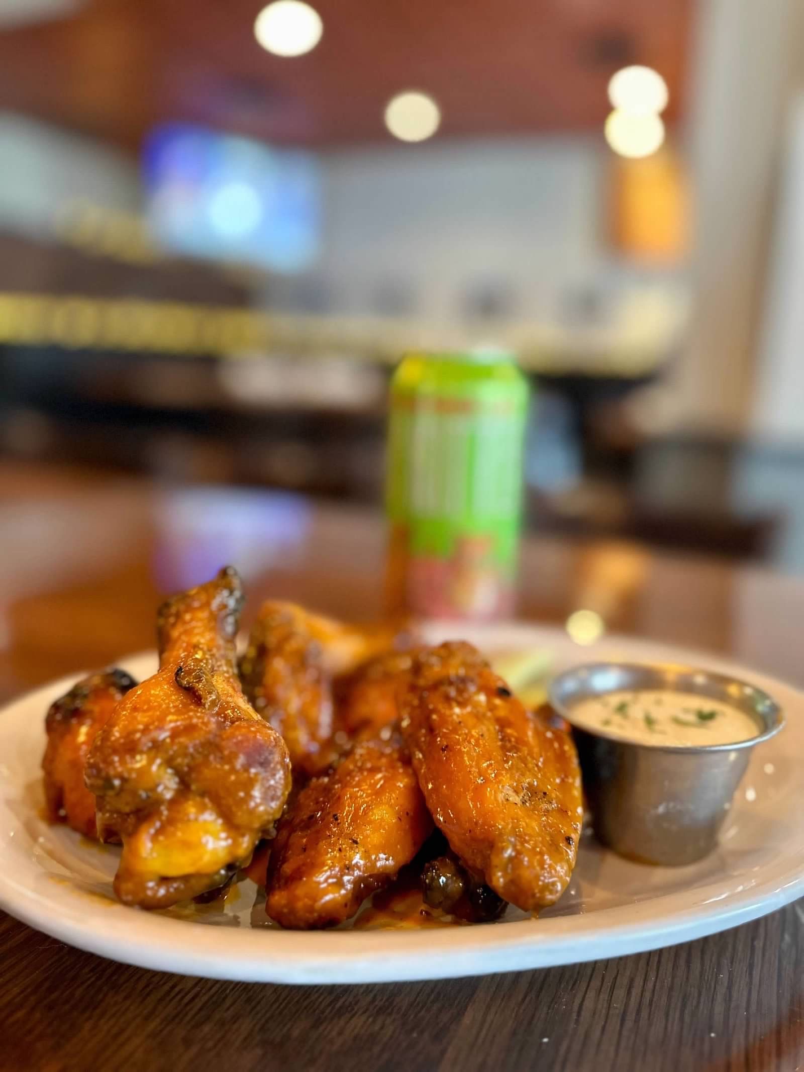 A plate of glazed chicken wings with a small cup of dipping sauce, with a blurred drink can in the background.