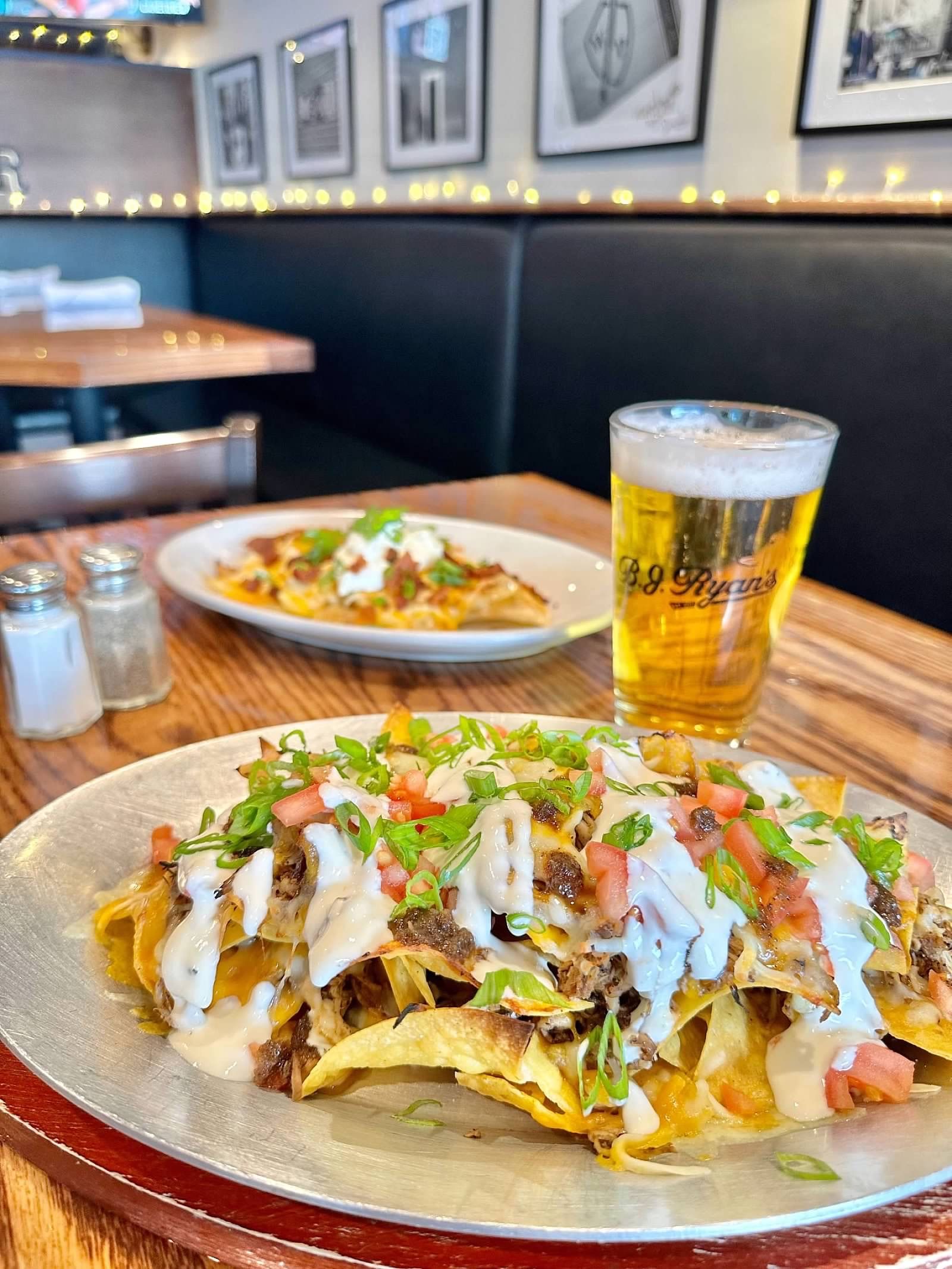A platter of nachos topped with sour cream, cheese, and scallions next to a glass of beer on a wooden table in a restaurant.