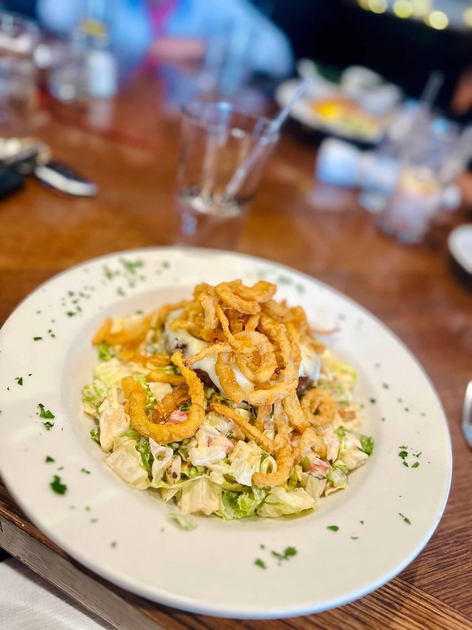 A close-up of a salad topped with crispy fried onion strings, served in a white bowl on a wooden restaurant table.