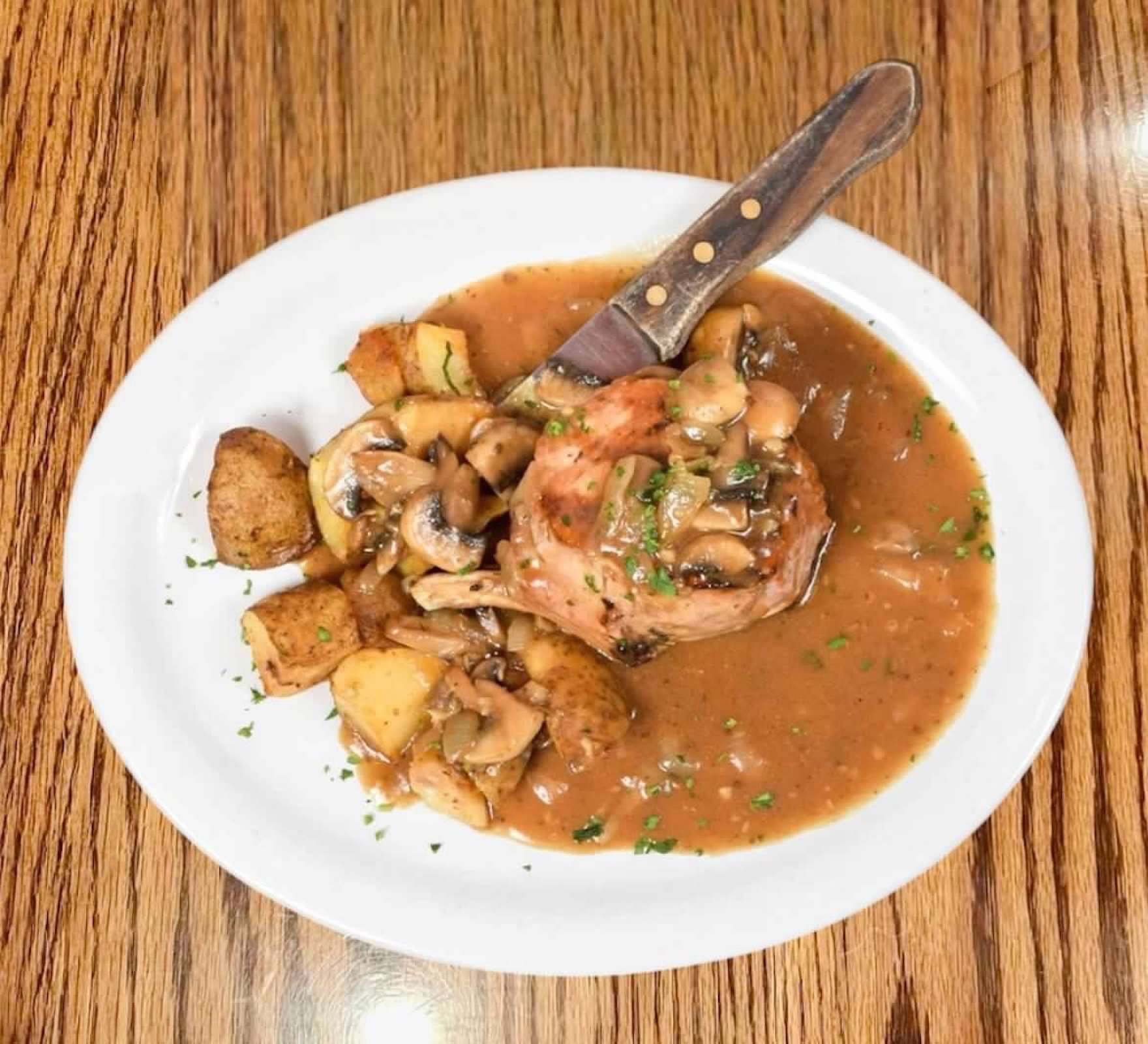 A plate featuring a meat portion, roasted potatoes, and mushrooms covered in a brown gravy with a knife resting on top.