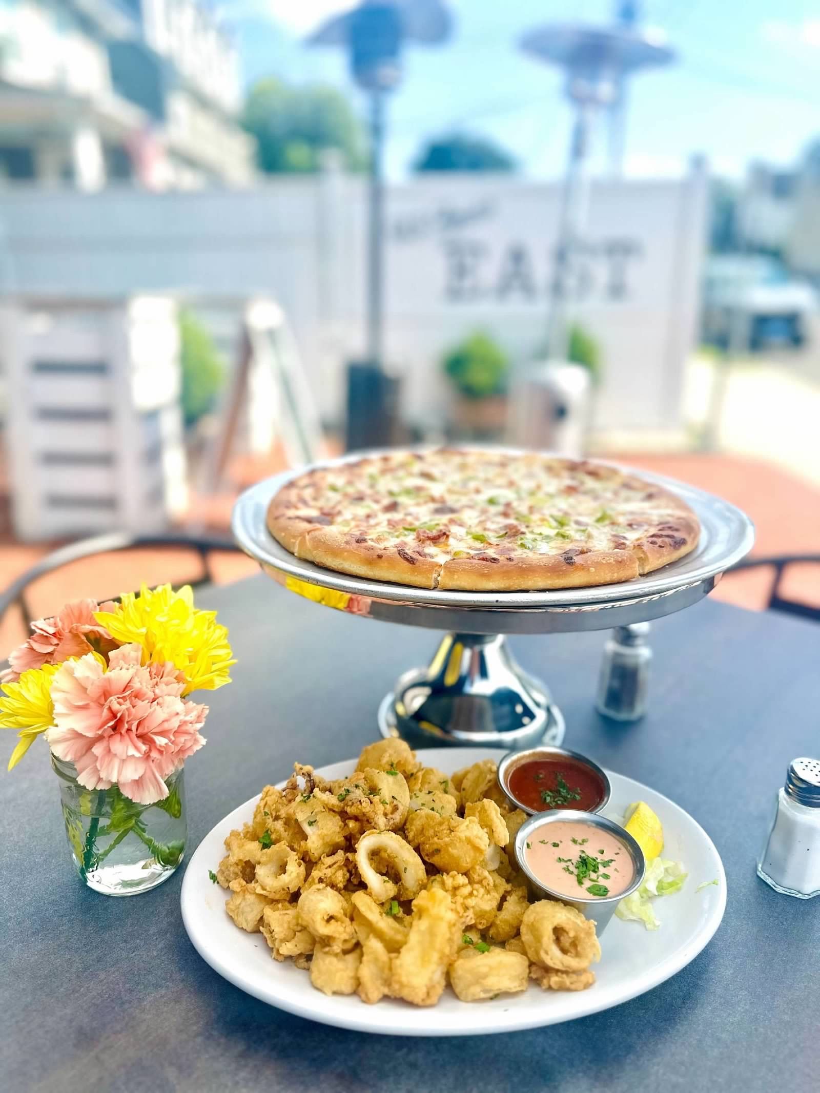 A pizza on a pedestal and a plate of fried calamari with two dipping sauces on a table outdoors next to flowers.