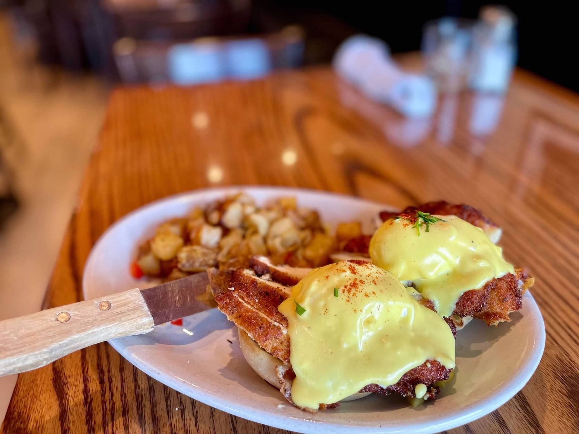 Two poached eggs on crispy chicken and English muffins, topped with hollandaise, served with potatoes on a wooden table.