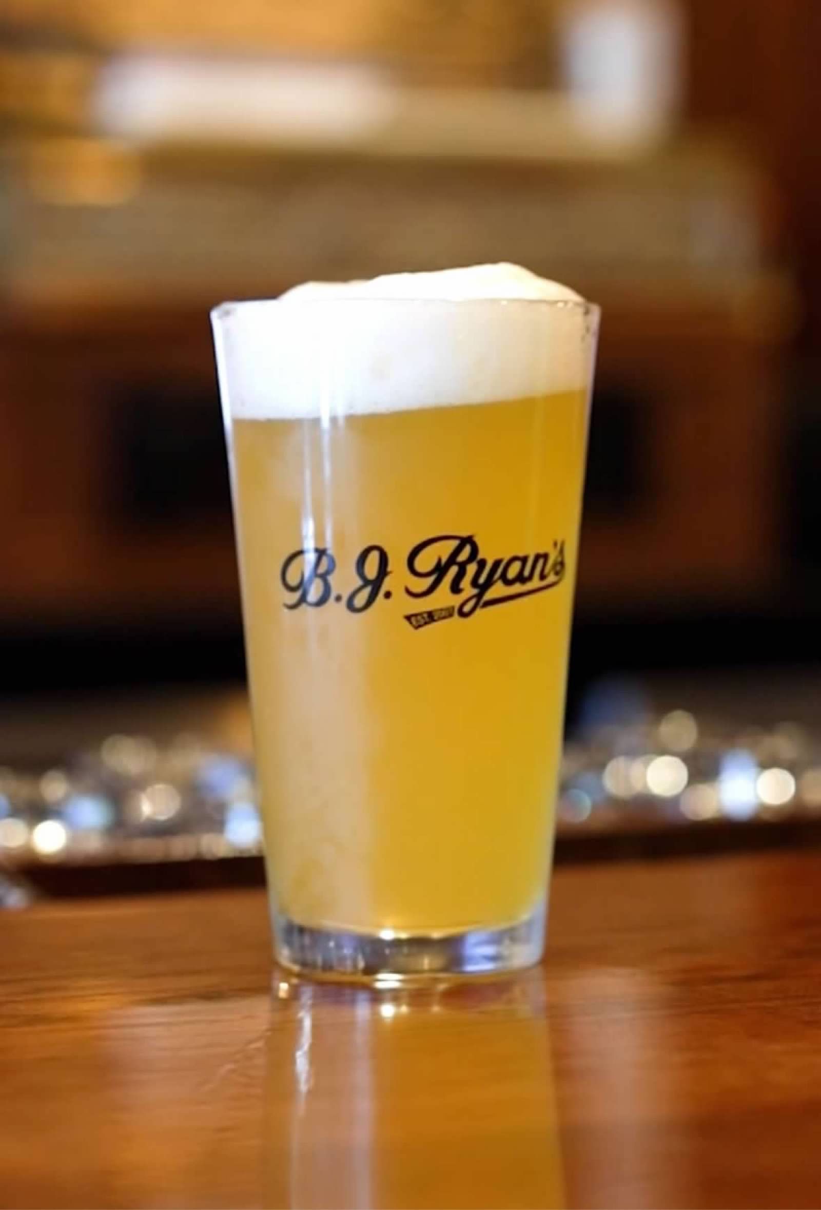 A glass of light-colored beer with a thick white head of foam, resting on a wooden bar top, featuring a B.J. Ryan's logo.