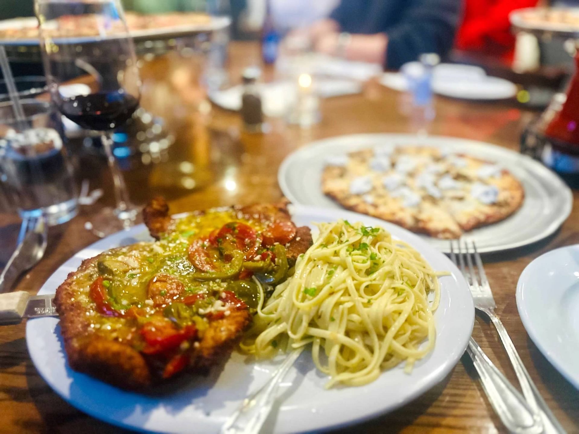 A breaded cutlet topped with peppers and a side of pasta sit on a table next to a pizza and a glass of wine.