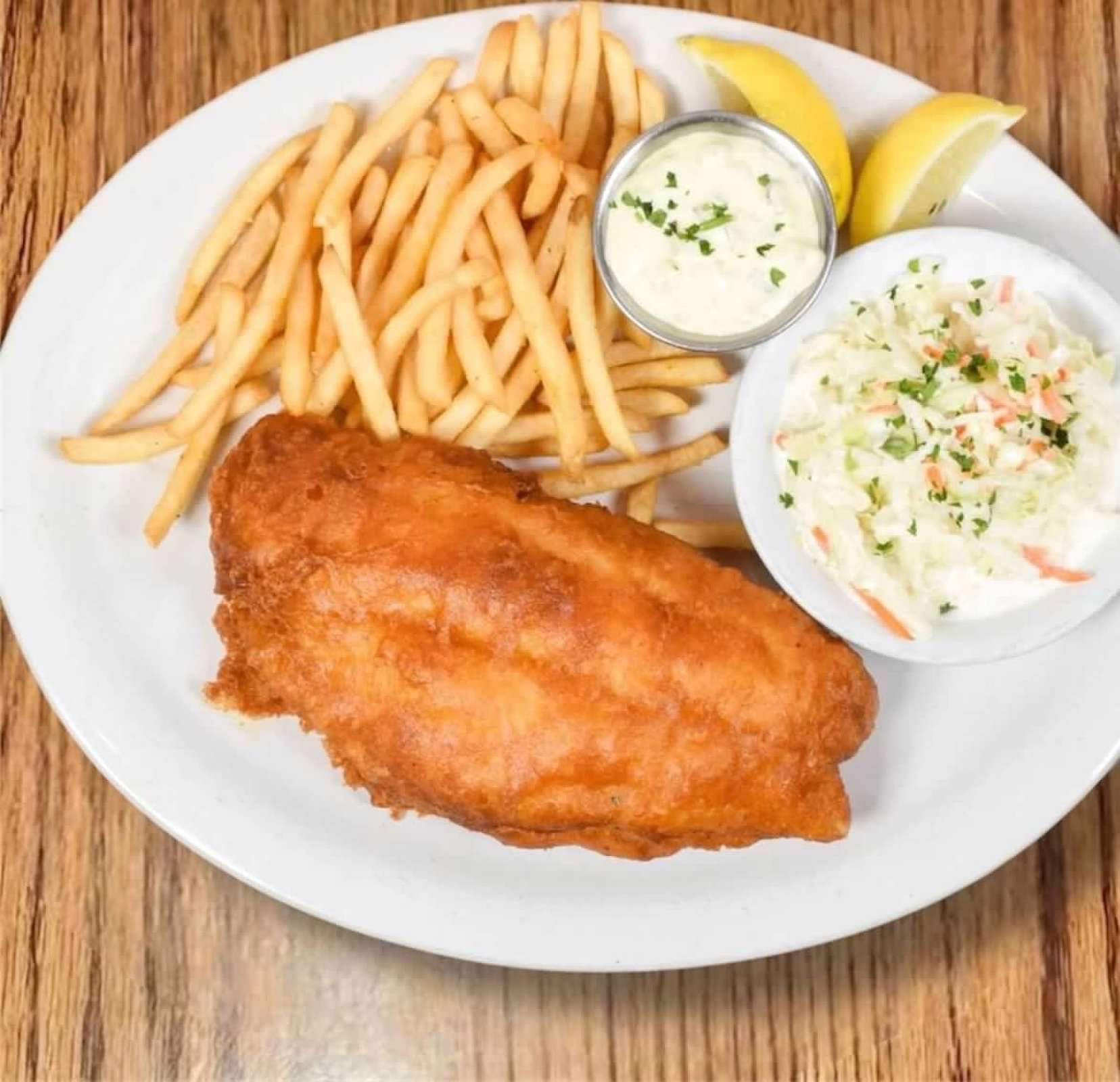A plate of golden-brown fried fish and thin-cut French fries, served with sides of coleslaw, tartar sauce, and lemon.
