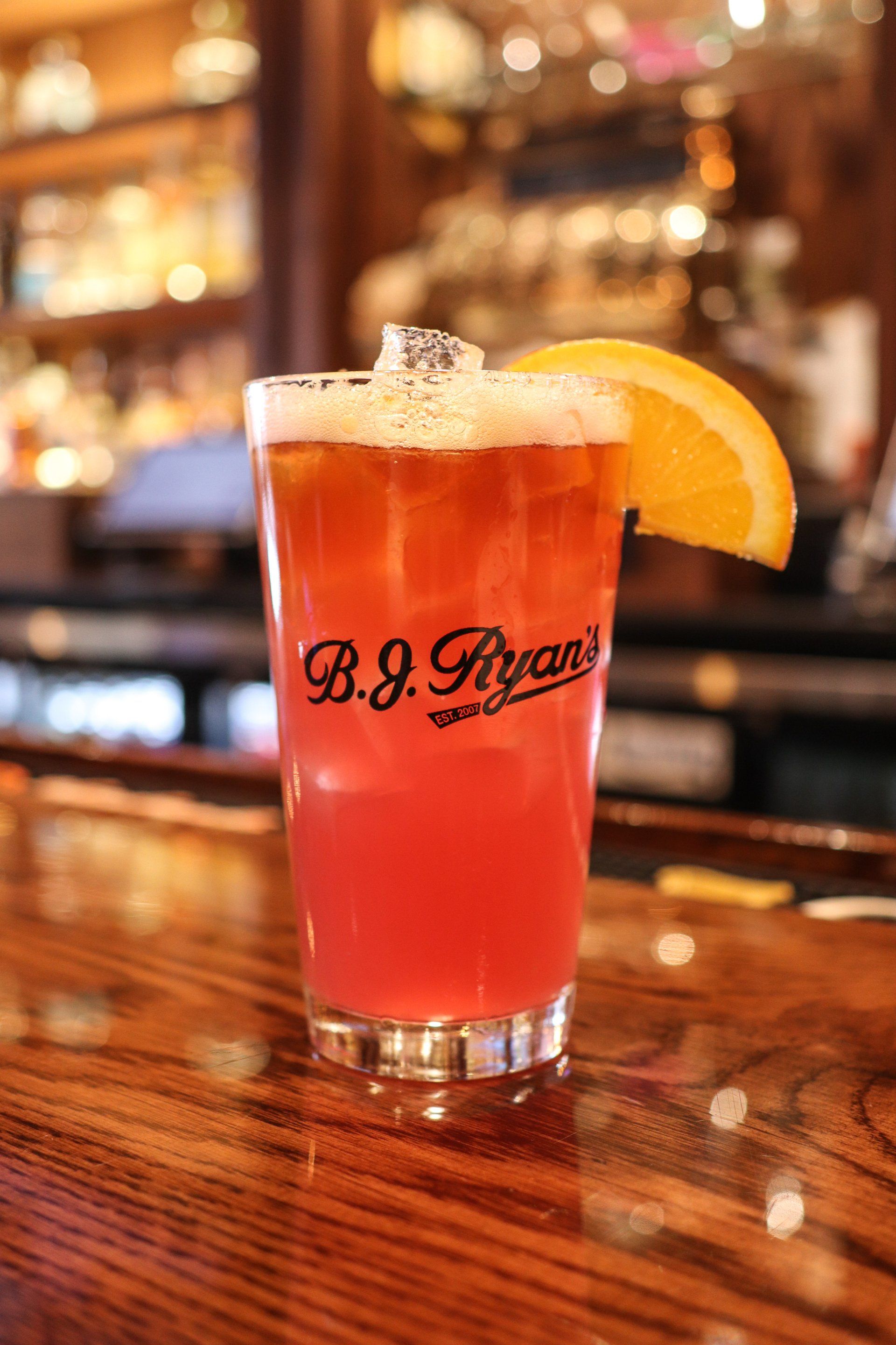 A glass of red cocktail with an orange slice garnish on a wooden bar top at B.J. Ryan’s, with a blurred pub background.