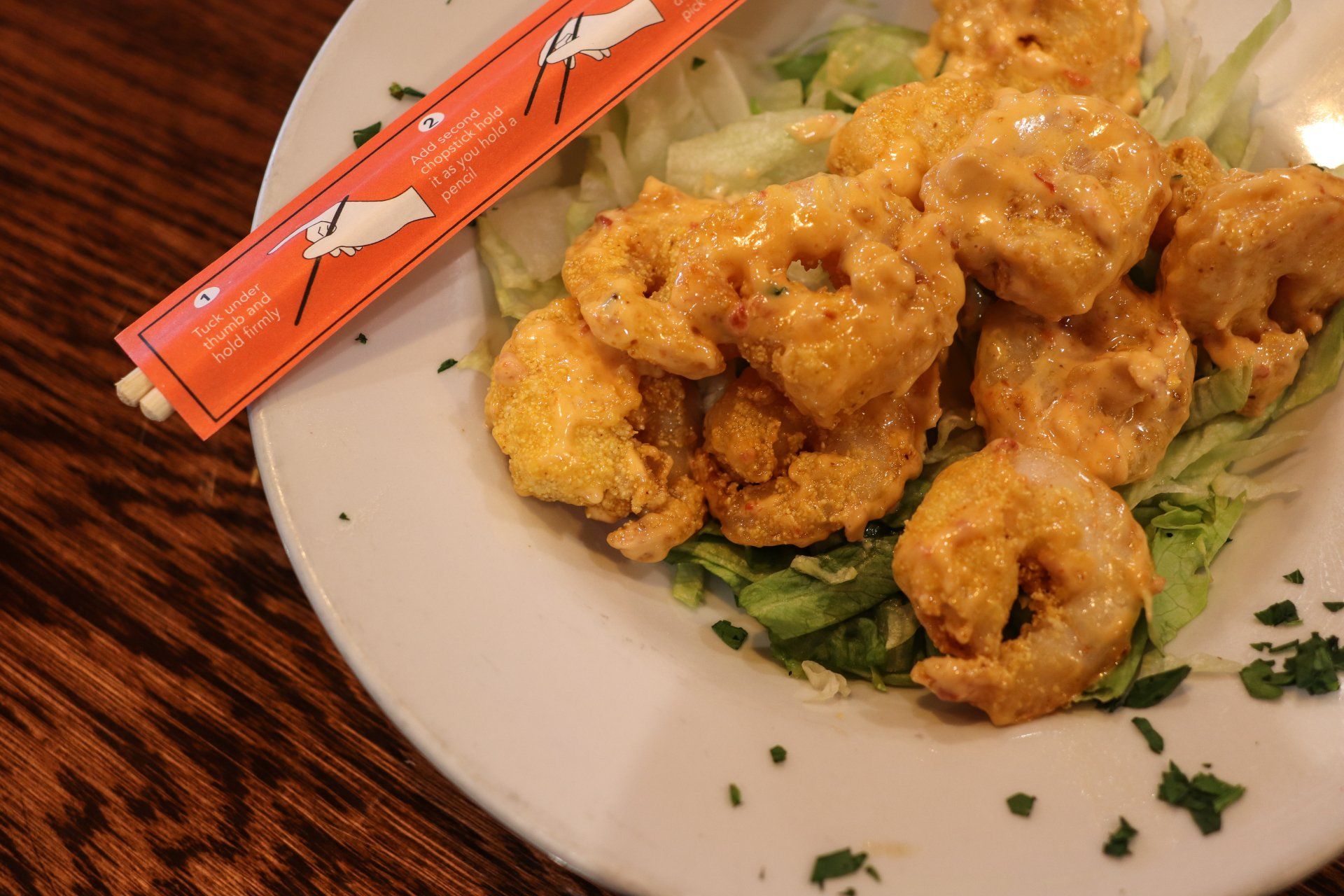A bowl of breaded, sauce-covered shrimp served over a bed of lettuce, with a chopstick wrapper resting on the side.