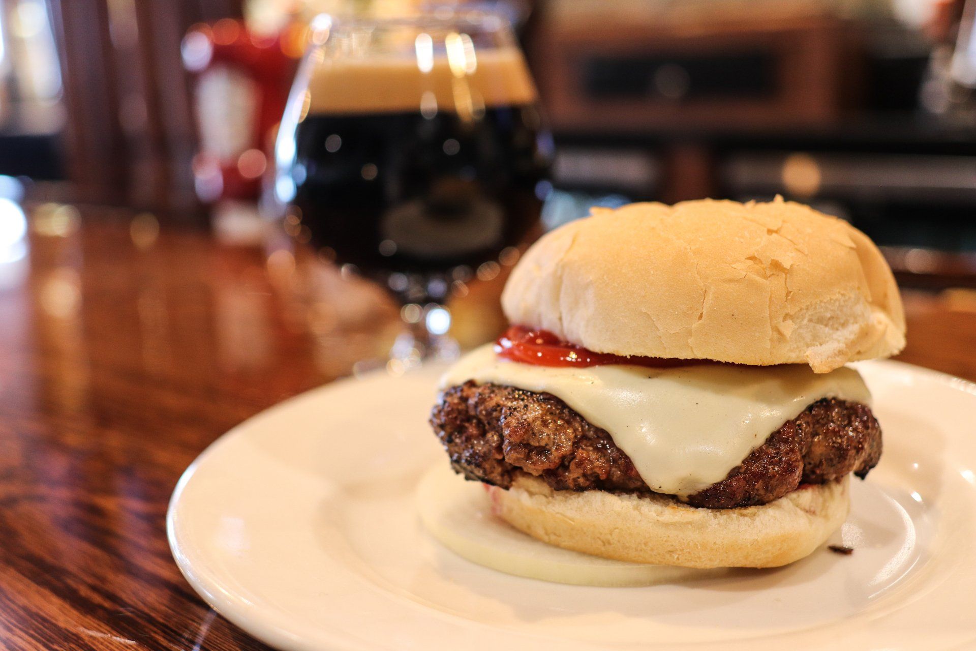 A cheeseburger with ketchup on a white plate, positioned in front of a glass of dark beer on a wooden bar counter.