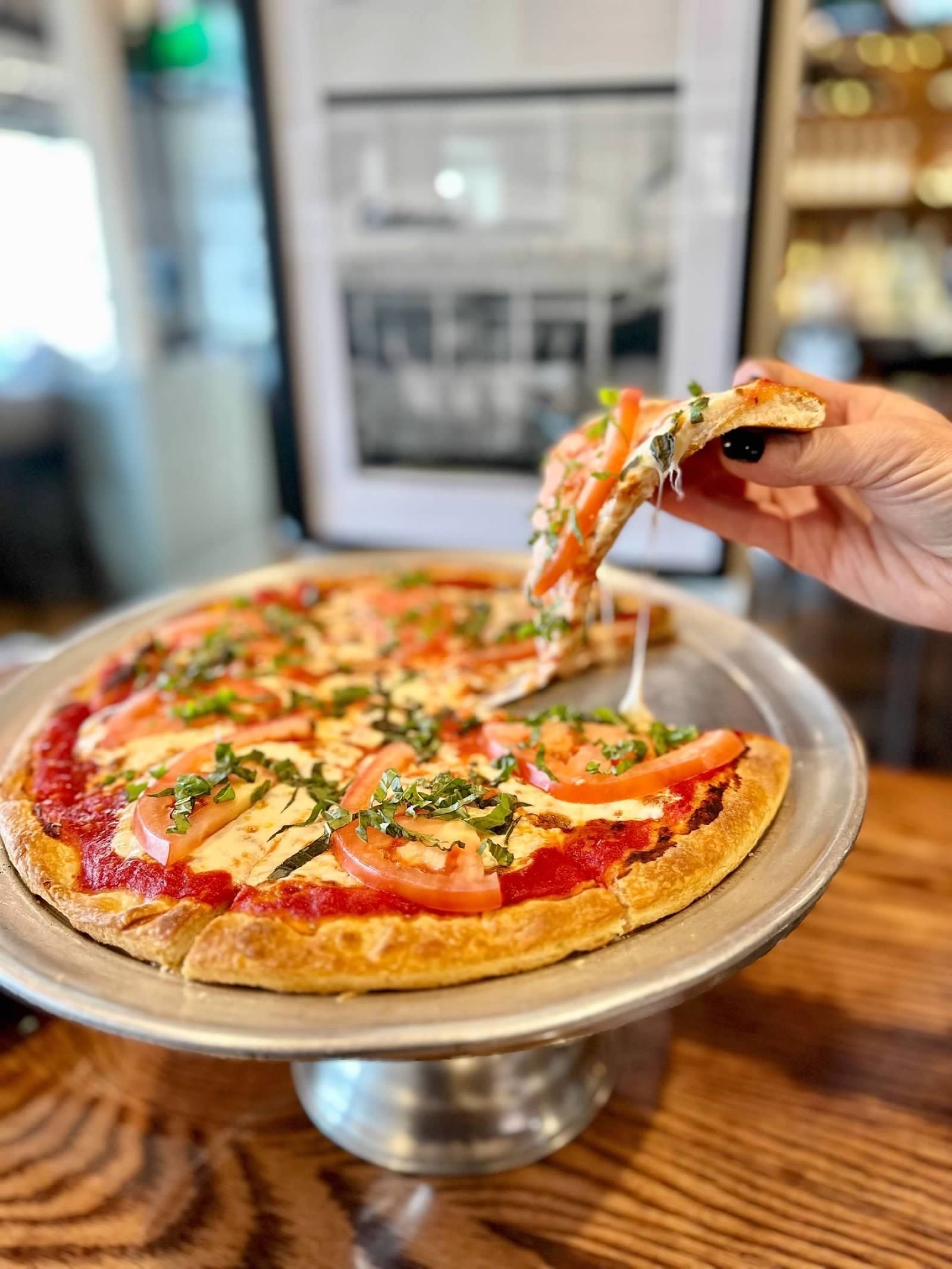 A hand lifts a slice of tomato and herb pizza from a metal stand in a restaurant setting.