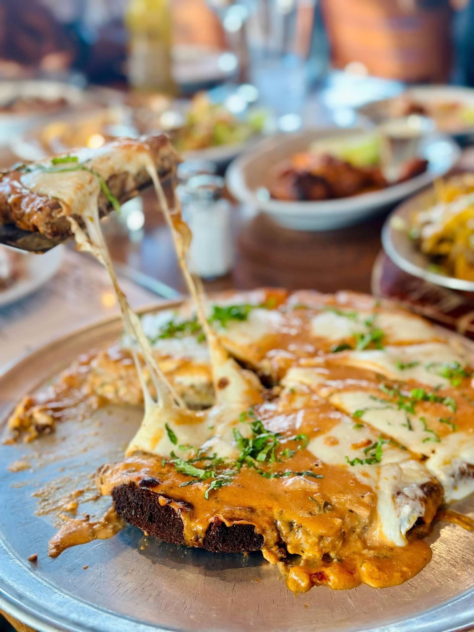 A pizza with stretchy melted cheese being lifted from a metal pan at a restaurant table.