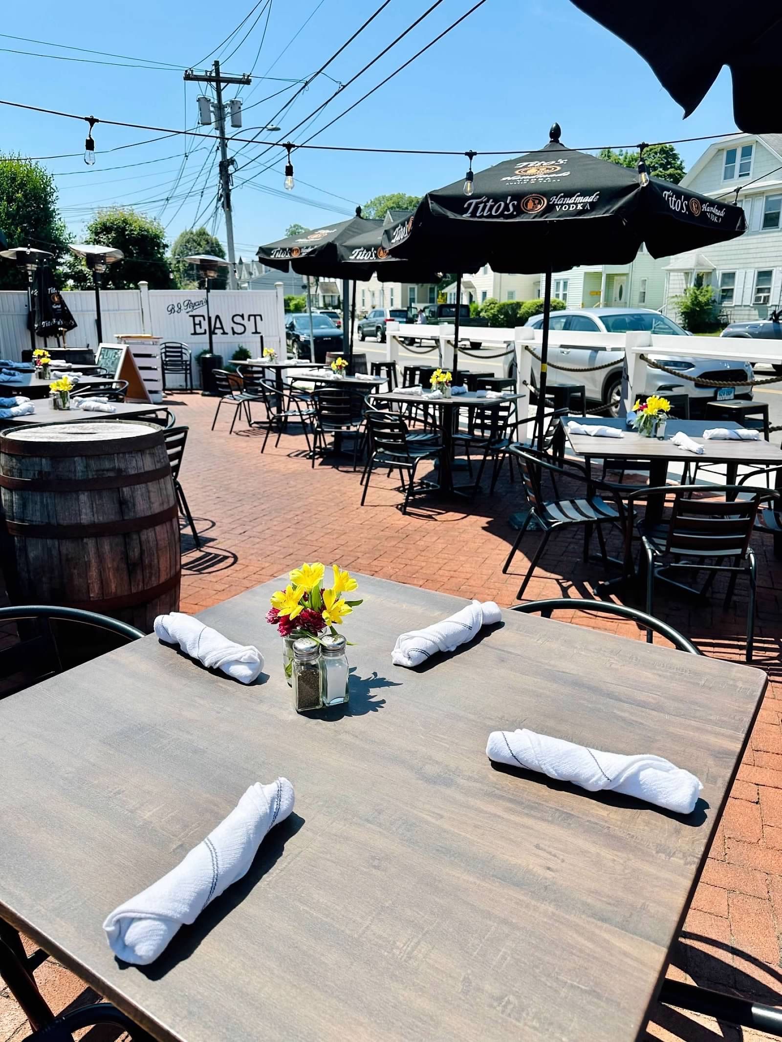 Outdoor patio dining area with multiple tables, black umbrellas, and string lights on a sunny day.
