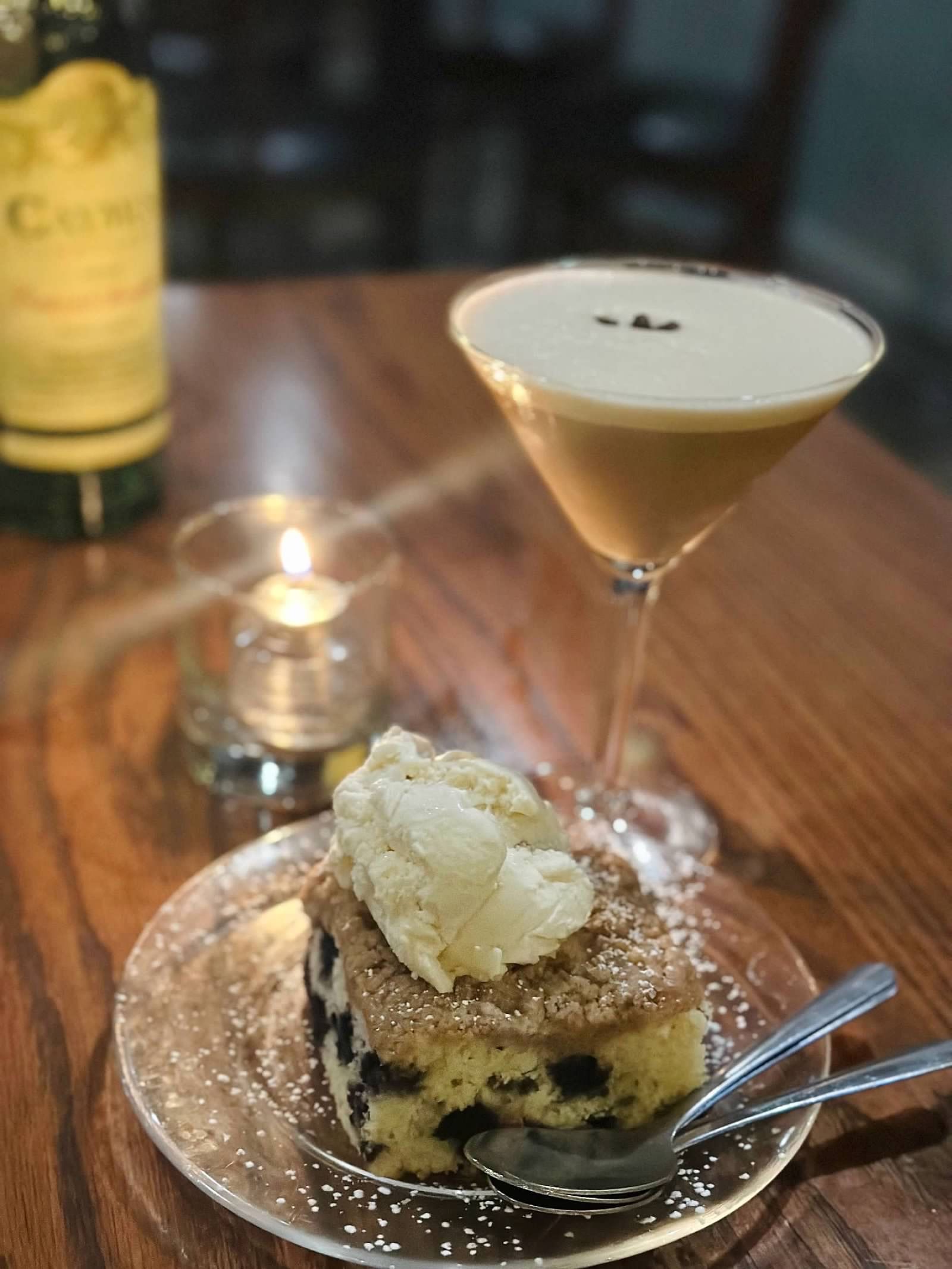 A slice of berry coffee cake topped with a scoop of vanilla ice cream, served with an espresso martini and a candle.