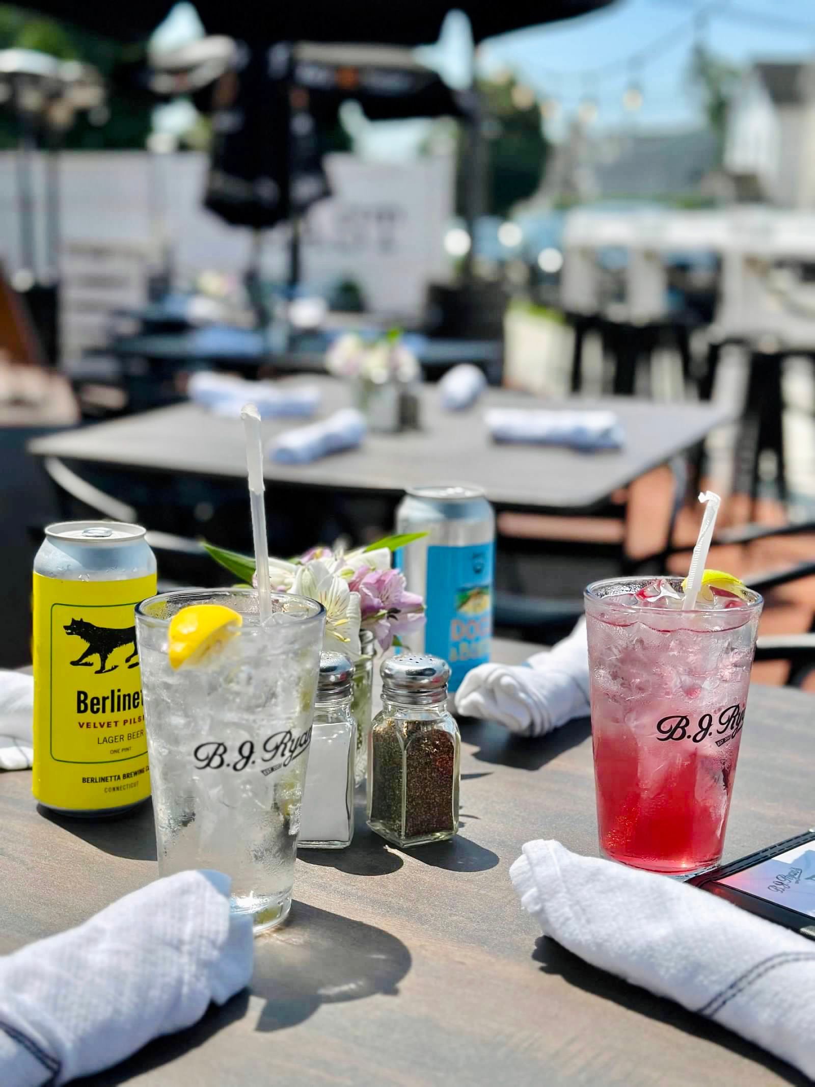 Outdoor restaurant table with drinks, a yellow can, seasonings, and white cloth napkins, with other tables blurred behind.