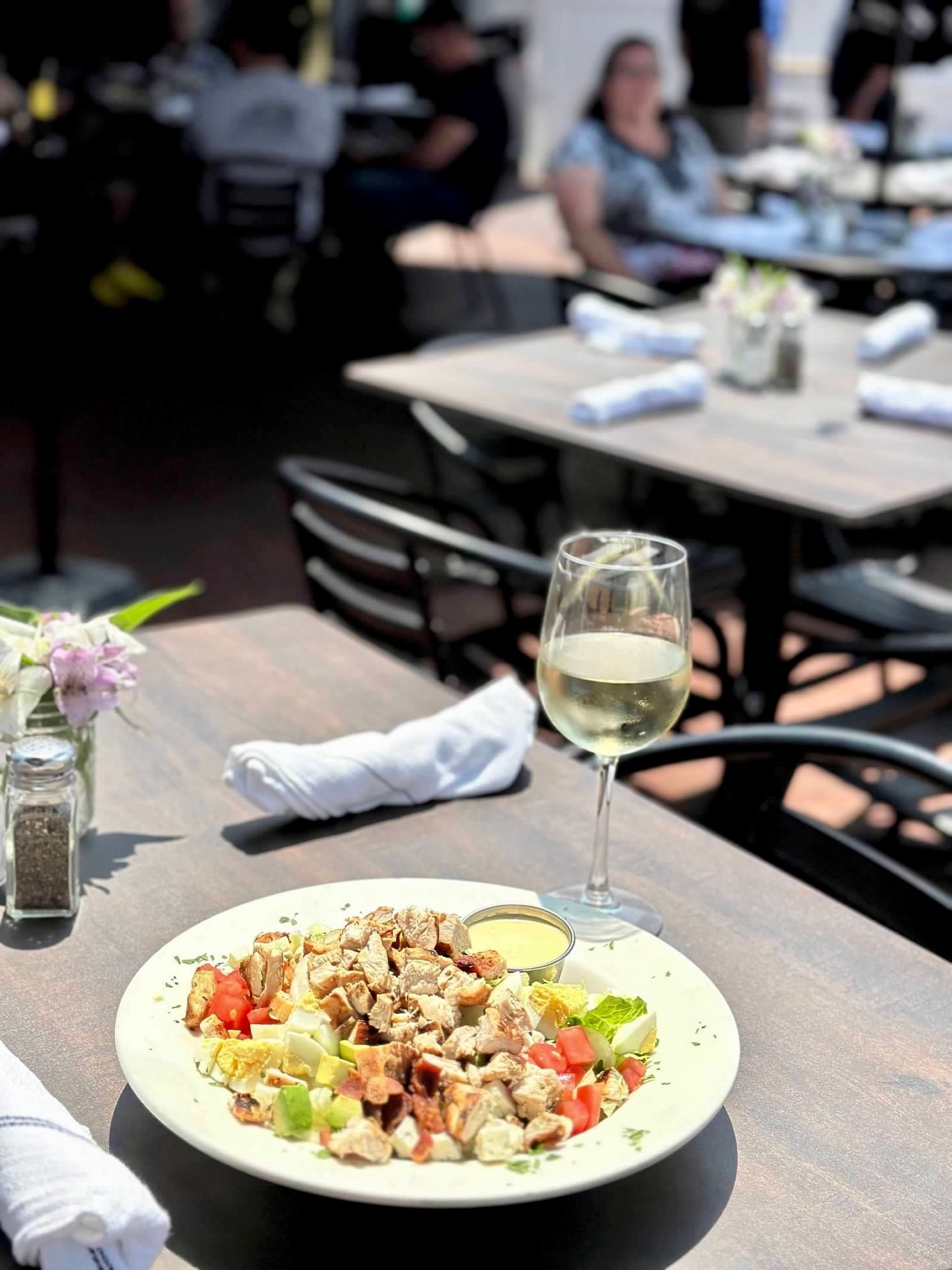A chicken salad and a glass of white wine on a restaurant table, with outdoor dining area in the background.
