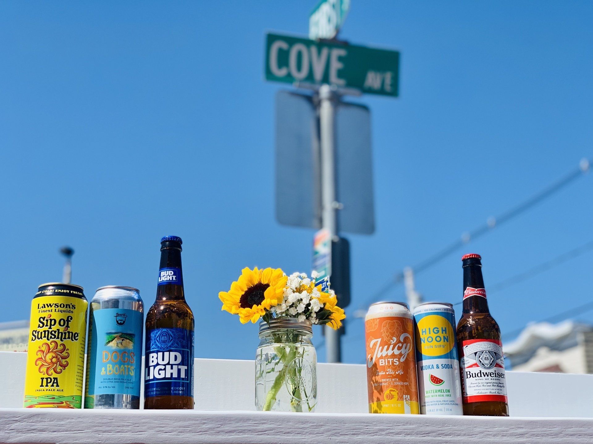 Six beer bottles and cans stand next to a mason jar of flowers in front of a street sign labeled 