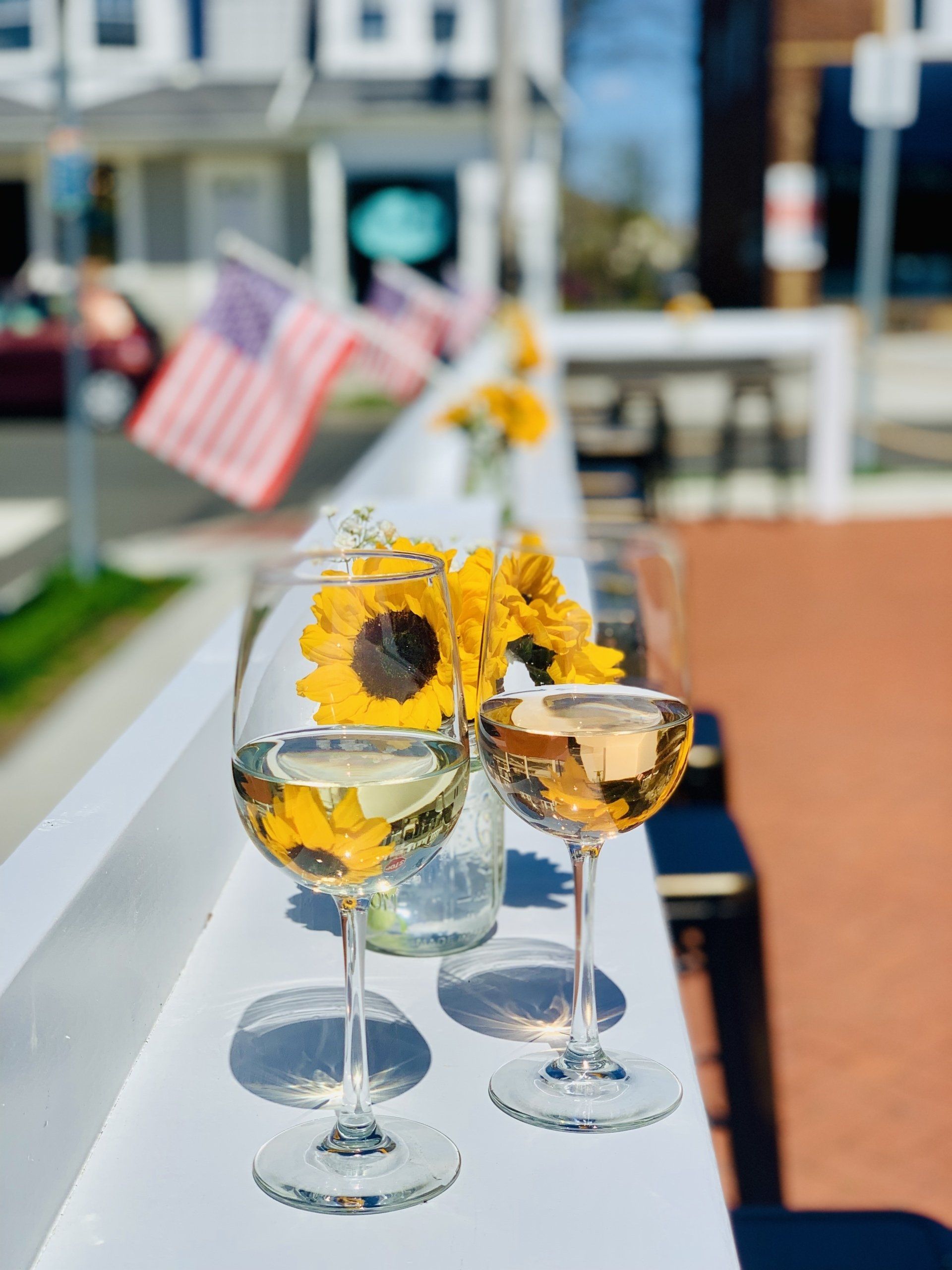 Two wine glasses and sunflowers on a white outdoor bar, with blurred American flags and street scenery in the background.