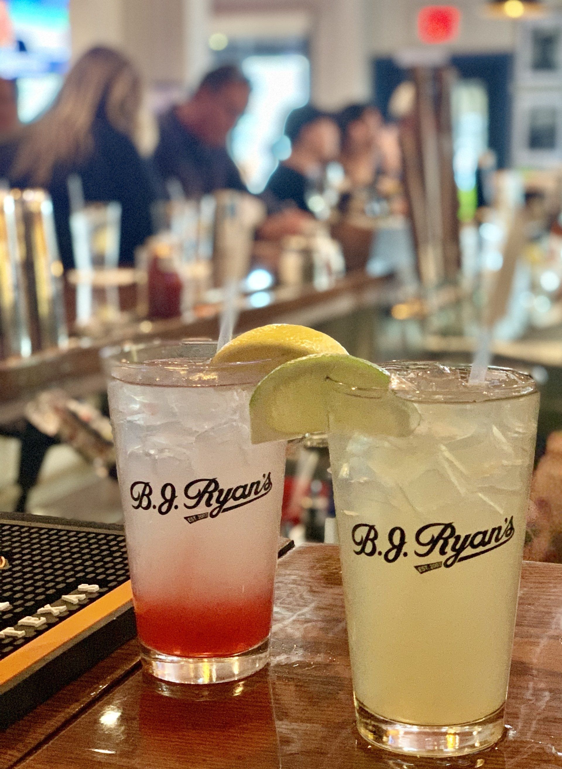 Two glasses with B.J. Ryan’s branding, one with a red-layered drink and one clear, sit on a bar counter with lemon garnishes.