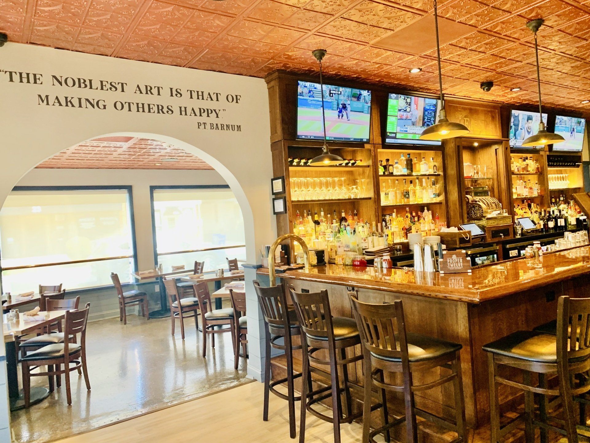 A bar interior with a copper ceiling, wooden counter, high stools, and an arched entryway leading to a dining area.