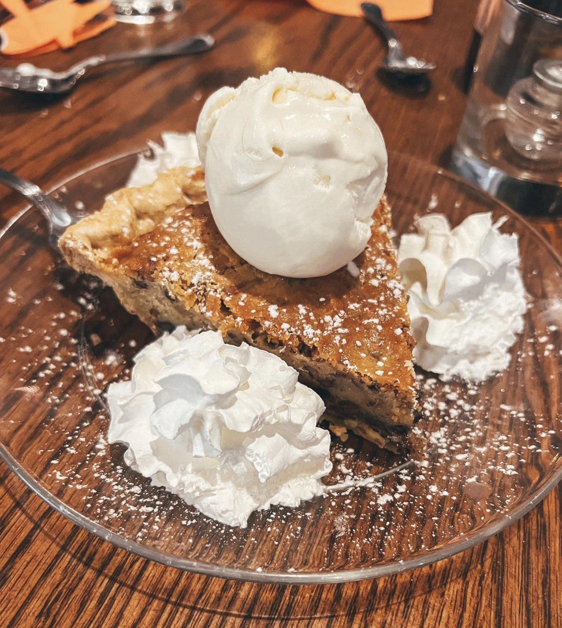 A slice of chocolate chip pie topped with a scoop of vanilla ice cream and two mounds of whipped cream on a clear plate.
